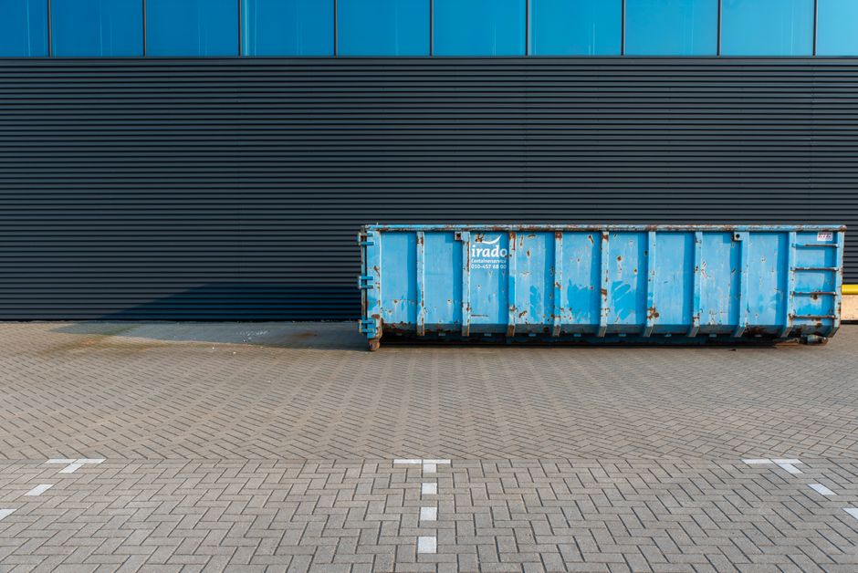 A large, weathered blue metal skip container positioned on a paved asphalt surface with a grid pattern, placed directly in front of a modern building with black horizontal cladding and a blue panel facade. The container, which shows signs of rust and paint chipping, features a white and blue logo for 'Irado' and a contact number, indicating it is used for waste or debris disposal during a home relocation or moving process. No people or objects are visible inside or around the skip. The scene is illuminated by natural daylight, casting a shadow of the container onto the pavement. This image illustrates the kind of waste management equipment that may be used for decluttering or disposal during furniture transport and packing activities associated with house removals, as handled by companies like Man With a Van Mitcham.