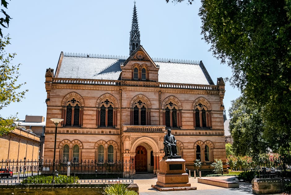 A large historic church building constructed from reddish-brown stone, featuring arched windows with decorative tracery and a central wooden door beneath a rounded archway. The church has a steep, slate-tiled roof and a tall, ornate spire extending into a clear blue sky. In front of the church, there is a paved pathway bordered by a black wrought iron fence, with a bronze statue of a seated figure on a pedestal situated on the sidewalk. The surrounding area includes green trees on the right and left sides, and a lamppost on the left. The scene appears to be captured during daylight hours with bright natural lighting, and the image context is consistent with a historic building near a residential area, which could be part of a move or relocation process, aligning with the services offered by Man With a Van Mitcham in house removals.