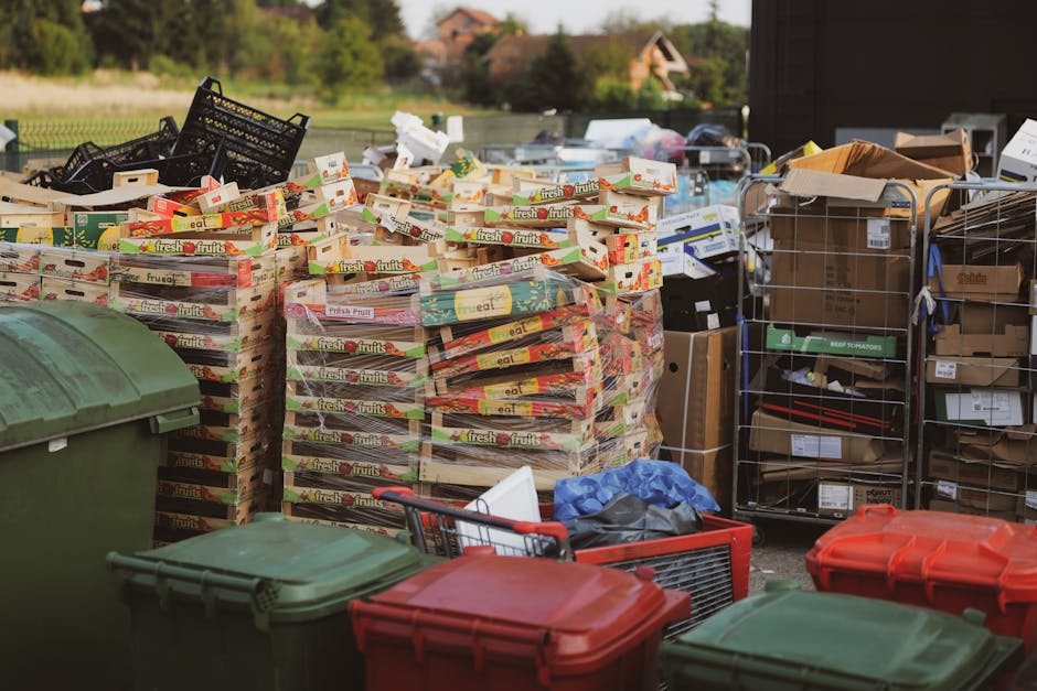 A large outdoor area near a property shows multiple stacks of flattened cardboard boxes labeled 'fresh fruits' and piled on wooden pallets, alongside metal cage containers filled with various boxes, some open, revealing packaging materials and miscellaneous household items. Several plastic trash bins in green and red are positioned in the foreground, while a collection of cardboard boxes and packaging materials are visible on a trolley and on the ground. The scene appears to be part of a home relocation or furniture transport process, with equipment such as pallets, trolleys, and packed boxes indicating loading or unloading activities associated with a professional removals service like Man With a Van Mitcham. The background includes a partially visible building and outdoor greenery, and the lighting suggests daytime with natural light illuminating the surroundings.