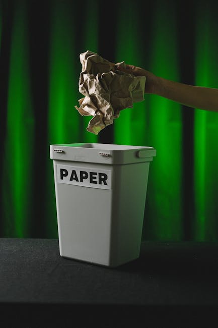A white paper recycling bin with a black label reading 'PAPER' positioned on a dark floor in front of a vibrant green curtain backdrop. An arm extends from the right side of the image, holding crumpled brown and white paper above the bin, about to dispose of it. The scene is illuminated with focused lighting, highlighting the stark contrast between the white bin and the vivid green background. This image illustrates waste disposal and recycling processes, which are integral to efficient home relocation and furniture transport services provided by Man With a Van Mitcham, including packing and loading during house removals.