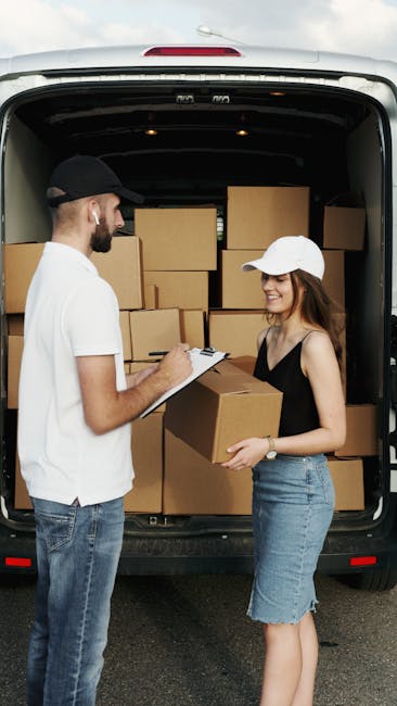 A man and a woman are standing outside the rear of a van used for house removals, with the van's back doors open to reveal an interior filled with stacked cardboard boxes of various sizes, some secured with plastic wrap. The man, wearing a black cap, white t-shirt, and jeans, is holding a clipboard and appears to be noting details related to the packing process. The woman, dressed in a white cap, black sleeveless top, and denim skirt, is smiling and holding a medium-sized cardboard box, ready to load it into the van. The scene is set outdoors on a paved surface, with natural daylight illuminating the area, and the environment suggests an organized packing and moving operation typical of home relocation services, as provided by Man With a Van Mitcham. The image captures the logistical aspect of furniture transport and packing in preparation for a house removal, reflecting effective load management and moving readiness.