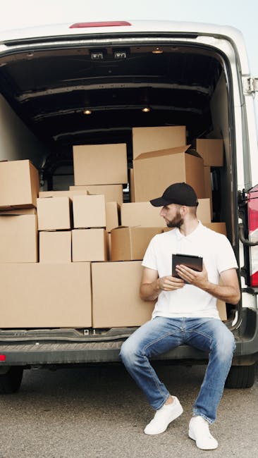 A man sitting on the ground next to an open van loaded with multiple cardboard boxes of various sizes, some sealed with packing tape, while others are open or partially filled. The man is wearing a black cap, white t-shirt, blue jeans, and white sneakers, and is holding a tablet device, possibly reviewing a moving plan. The van is parked on a paved surface, with the interior showing a dark cargo space filled with packed boxes arranged for safe transport during a home relocation. The scene illustrates the packing and loading process associated with professional removals services, such as those offered by Man With a Van Mitcham, emphasizing careful handling and organized transportation of household items.
