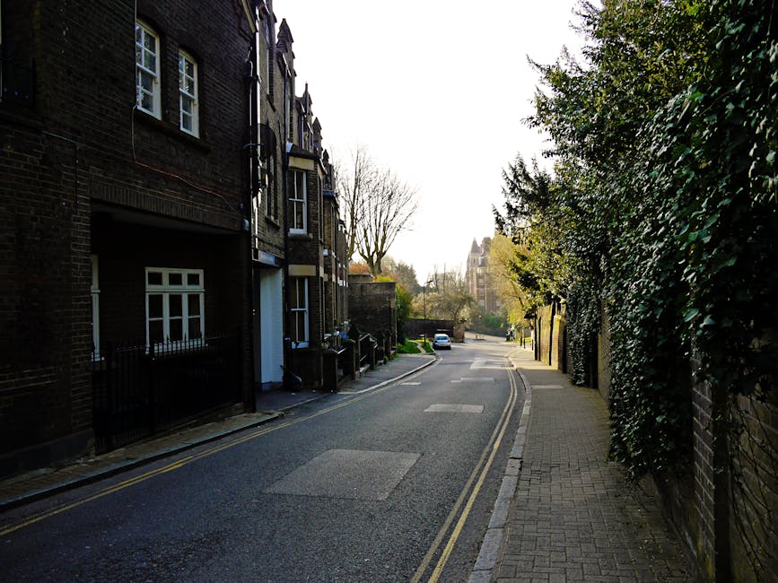 A narrow residential street with a slight curve, featuring a pavement on the right side lined with lush green and dark foliage, and a row of brick terraced houses on the left. The houses have large windows with white frames and some have black iron railings at the front. A white van or moving truck belonging to Man With a Van Mitcham is partially visible on the left, parked close to the residences, with the loading area near the house entrance. The street appears quiet with minimal traffic, and a single car is seen further in the distance. The scene is well-lit with natural daylight, and leafless trees are visible in the background, indicating early spring or late autumn. This setting depicts a typical UK street suitable for home relocation and furniture transport services, with clear outdoor space for loading or unloading boxes, furniture, and appliances during a house move.