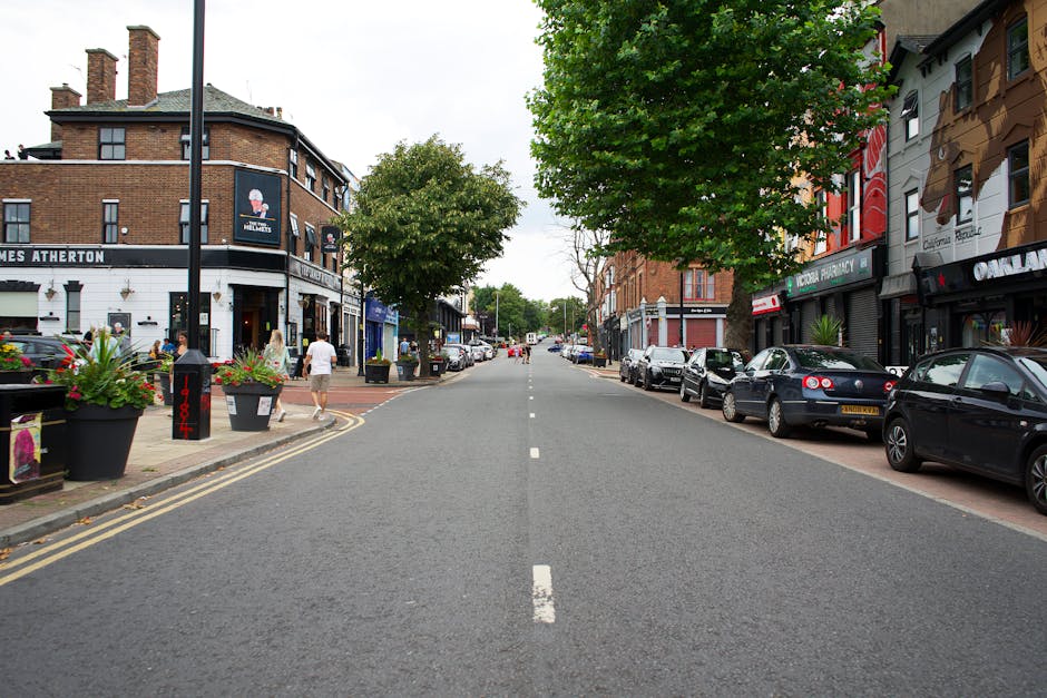 A straight section of London Road in Mitcham lined with parked cars on both sides, including sedans and hatchbacks, with a few pedestrians walking along the sidewalk. On the left, there is a building with a white facade housing a pub or restaurant, identifiable by outdoor seating and flower planters. On the right, there are various commercial storefronts with signage, some of which display images or logos, and a row of bungalows or low-rise buildings with residential windows. Several mature green trees with full foliage are evenly spaced along the street, casting partial shade. Street elements include black lampposts, a black bollard with red markings, and double yellow lines along the curb indicating parking restrictions. The road surface is clean and well-maintained, featuring a dashed white line down the center, and the surrounding ambient lighting suggests daytime with overcast skies. The scene captures a typical urban setting suitable for house removals or furniture transport, highlighting the street environment where [COMPANY_NAME] may conduct moving or packing services as part of an upcoming home relocation process.