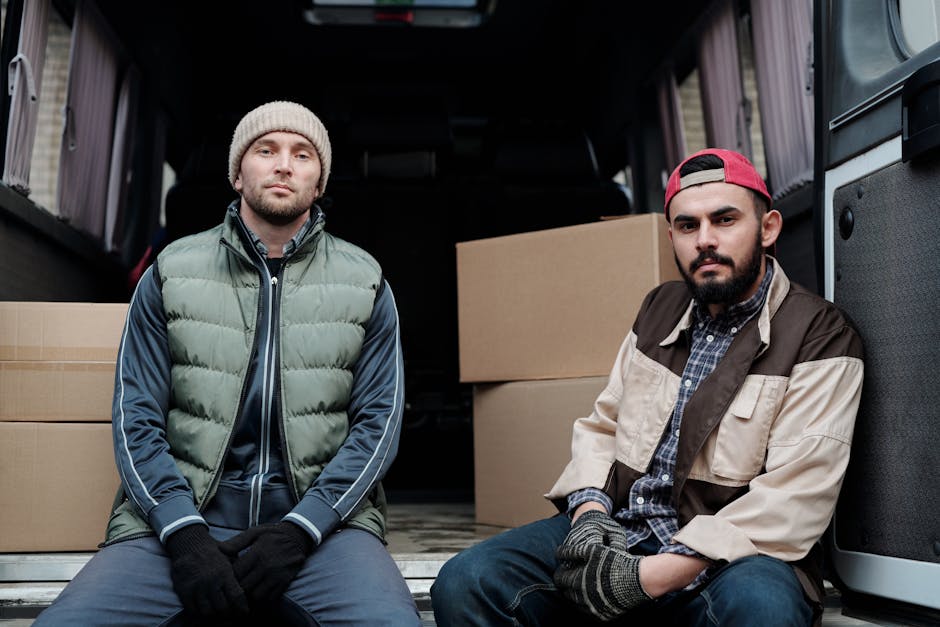 Two professional movers wearing dark uniforms are loading a large, green-draped piece of furniture into the open cargo area of a black van parked on a street. The van is positioned with its rear doors