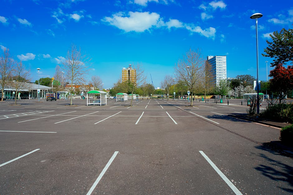 A large, nearly empty parking lot in Mitcham under a bright blue sky with scattered clouds. The asphalt surface is marked with white lines designating individual parking spaces, with some spaces containing small, white, shelter-like structures providing shade. Surrounding the lot are a few leafless and some blossoming trees, along with low-rise buildings and taller office blocks in the background. A single lamppost stands on the right side, casting a shadow on the pavement. The overall scene depicts a quiet, well-organized area suitable for home relocation or moving vehicles, with no cars currently parked, and the environment appears clean and maintained. The image subtly supports themes related to removals and furniture transport, aligning with house moving services provided by [COMPANY_NAME].