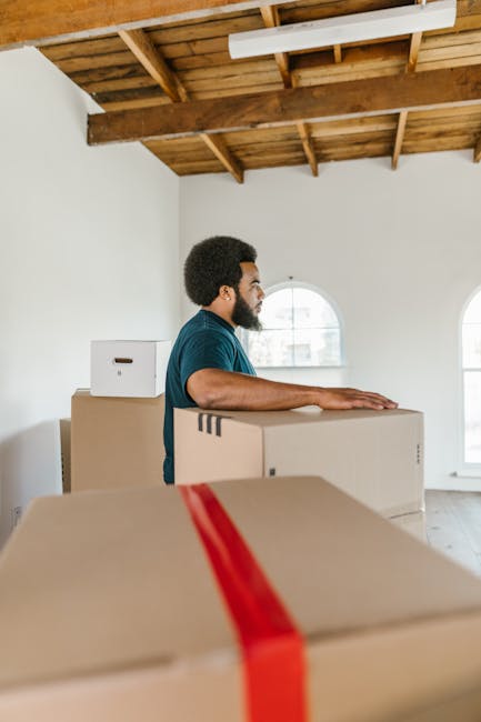 A man with dark curly hair and a beard, dressed in a blue T-shirt, is standing inside a room with white walls and a wooden ceiling, next to a stack of packed cardboard boxes. He is resting his hands on one of the boxes, which is secured with red packing tape, preparing for a home relocation or furniture transport. The room has large, arched windows allowing natural light to illuminate the space. In the background, more boxes are visible, indicating an ongoing packing process essential for professional removals. The environment appears clean and organized, reflecting a careful packing and loading process typical of house moving services offered by Man With a Van Mitcham. The scene captures the early stages of loading items into a vehicle for transportation, with the focus on safe handling and efficient logistics involved in residential moves.