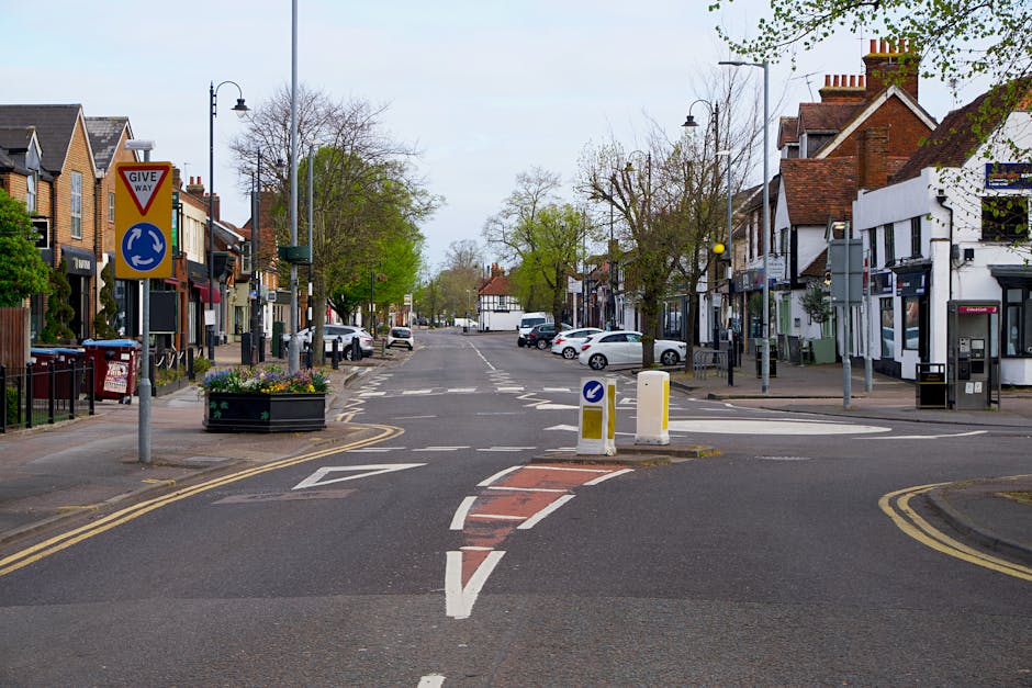 An urban residential street scene showing a clear view of a small roundabout with a 'Give Way' sign on the left side and a blue directional arrow sign indicating traffic flow. The roadway includes designated lanes marked with dashed white lines and a painted red and white warning zone for vehicles. On either side of the street, there are brick and white-painted shop-front buildings, some with awnings, and parked cars lining the pavements. Leafless trees and some with light green foliage are visible, suggesting early spring or late autumn, with overcast sky lighting the scene evenly. Limited pedestrian activity is present, with street lighting fixtures and a small planter box with flowers positioned near the roundabout. This setting provides a typical backdrop for house removals or local moving services, illustrating street access that can impact packing and furniture transport logistics, as would be considered by Man With a Van Mitcham during home relocations.