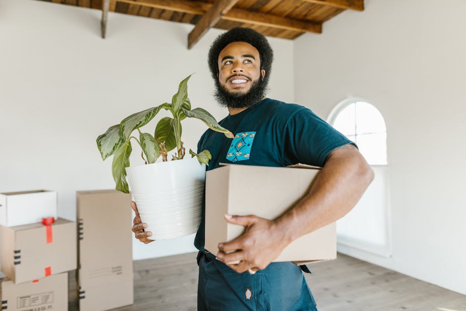 A man with dark curly hair and a beard, dressed in a blue T-shirt, is standing inside a room with white walls and a wooden ceiling, next to a stack of packed cardboard boxes. He is resting his hands on one of the boxes, which is secured with red packing tape, preparing for a home relocation or furniture transport. The room has large, arched windows allowing natural light to illuminate the space. In the background, more boxes are visible, indicating an ongoing packing process essential for professional removals. The environment appears clean and organized, reflecting a careful packing and loading process typical of house moving services offered by Man With a Van Mitcham. The scene captures the early stages of loading items into a vehicle for transportation, with the focus on safe handling and efficient logistics involved in residential moves.