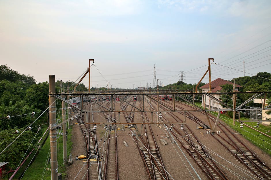 A wide-angle view of multiple railway tracks converging and diverging, with overhead electrical wiring supported by tall wooden and metal poles, situated in a semi-urban environment with green foliage on either side. The tracks are made of steel rails fixed on wooden sleepers, with gravel ballast underneath, extending into the distance beneath a partly cloudy sky. In the background, several red and white train carriages are visible stationary or moving along the tracks. To the right, there is a small building with a sloped roof and a green grassy area, indicating proximity to residential or office premises. Overhead electrical wires are strung between supporting poles, used for train power supply, and the scene is typical of a busy rail junction or station area. This image, associated with house removals and relocation services, captures the logistics of freight or passenger transport infrastructure which often complements home relocation processes managed by companies like Man With a Van Mitcham.