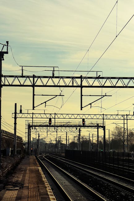 A view of railway tracks at a station during daylight, with overhead electric lines supported by metal structures. The tracks curve gently to the right, leading towards the horizon where the sky appears partly cloudy with soft, diffuse lighting. On the left side, a platform with tactile paving is visible, with some scattered objects possibly including small boxes or bags. In the background, there are trees and railway signals, indicating an active rail transport environment. The image provides a clear perspective of rail infrastructure that could be relevant to house removals and moving logistics when transferring belongings via train links, supporting the services offered by Man With a Van Mitcham for home relocation, furniture transport, and packing and moving operations.
