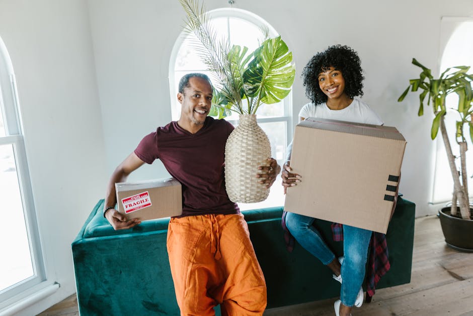 A middle-aged man and a young woman are indoors, smiling while holding packing and moving supplies during a home relocation. The man, wearing a maroon t-shirt and orange trousers, is holding a small cardboard box labeled 'Fragile' in one hand and a large woven vase with green foliage in the other. The woman, dressed in a white top and blue jeans with a red plaid shirt tied around her waist, is holding a medium-sized cardboard box. They are seated on a teal sofa positioned near a window with white trim, allowing natural daylight to illuminate the room. Behind them, there is a large potted plant with broad green leaves and a white wall, indicating an interior space prepared for the packing and moving process. The image captures the early stages of furniture transport and packing for a house move, representing professional removals services by Man With a Van Mitcham, supporting efficient and careful home relocations.