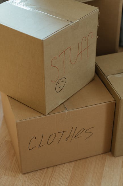 Close-up image of two brown cardboard boxes placed on a wooden floor, one stacked on top of the other. The top box has the word 'STUFF' written in red marker along with a simple smiley face, while the lower box is labeled 'CLOTHES' in similar handwriting. Both boxes appear to be sealed with packing tape and are used for packing personal belongings during a home relocation. The setting suggests an indoor environment, likely a room being prepared for moving or packing, with additional boxes partially visible nearby. The image reflects typical steps involved in furniture transport and packing during a house removal process, with a focus on organized packing materials managed by professional removal services such as Man With a Van Mitcham.