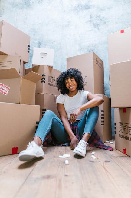 A middle-aged man and a young woman are indoors, smiling while holding packing and moving supplies during a home relocation. The man, wearing a maroon t-shirt and orange trousers, is holding a small cardboard box labeled 'Fragile' in one hand and a large woven vase with green foliage in the other. The woman, dressed in a white top and blue jeans with a red plaid shirt tied around her waist, is holding a medium-sized cardboard box. They are seated on a teal sofa positioned near a window with white trim, allowing natural daylight to illuminate the room. Behind them, there is a large potted plant with broad green leaves and a white wall, indicating an interior space prepared for the packing and moving process. The image captures the early stages of furniture transport and packing for a house move, representing professional removals services by Man With a Van Mitcham, supporting efficient and careful home relocations.