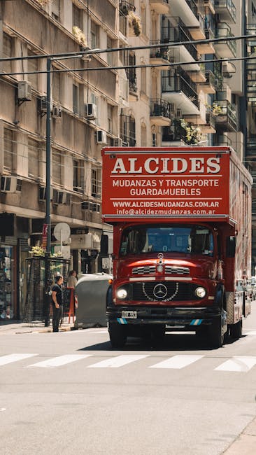 A bright red moving truck with the branding 'ALCIDES' and contact information parked on a city street, adjacent to multi-storey residential buildings with balconies, some with potted plants. The truck is positioned near the curb, with the front facing the camera, and appears to be involved in a home relocation or furniture transport process. The street scene includes pedestrians walking on the sidewalk and a person standing near a parked bicycle. The environment suggests an urban setting with typical city architecture, and the truck is likely preparing for or engaged in house removals or moving services, as offered by companies like Man With a Van Mitcham. The image captures the loading or unloading area, with elements like the truck's rear doors possibly open for loading furniture or boxes, and street lighting and shadows visible in natural daylight.